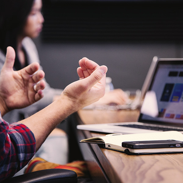 Man sitting in front of a laptop and notebook with his hands gesturing in conversation.
