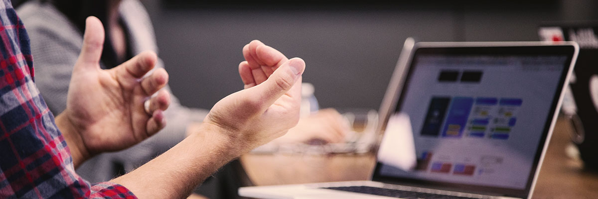 Man sitting in front of a laptop and notebook with his hands gesturing in conversation.